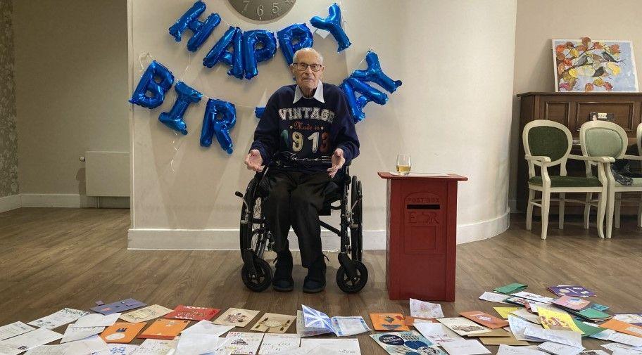 A man in a blue jumper which says "vintage 1913" sits in a wheelchair in front of rows of birthday cards on the floor, with blue balloons spelling "Happy Birthday" on the wall