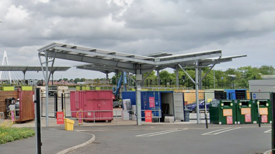 The entrance to Pallion Household Waste and Recycling Centre in Sunderland. There are a number of red, blue and green shipping containers in different sizes, surrounded by parking spaces.  A row of green trees is visible behind the facility, with the Northern Spire bridge further back.