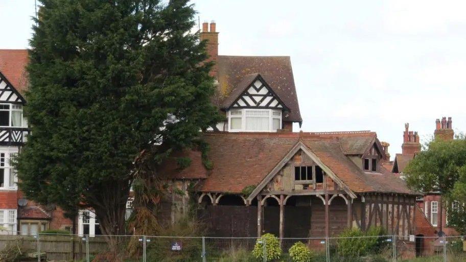 A ramshackle Victorian building, flanked by trees and surrounded by a metal fence. Some Tudor-style houses loom in the background.