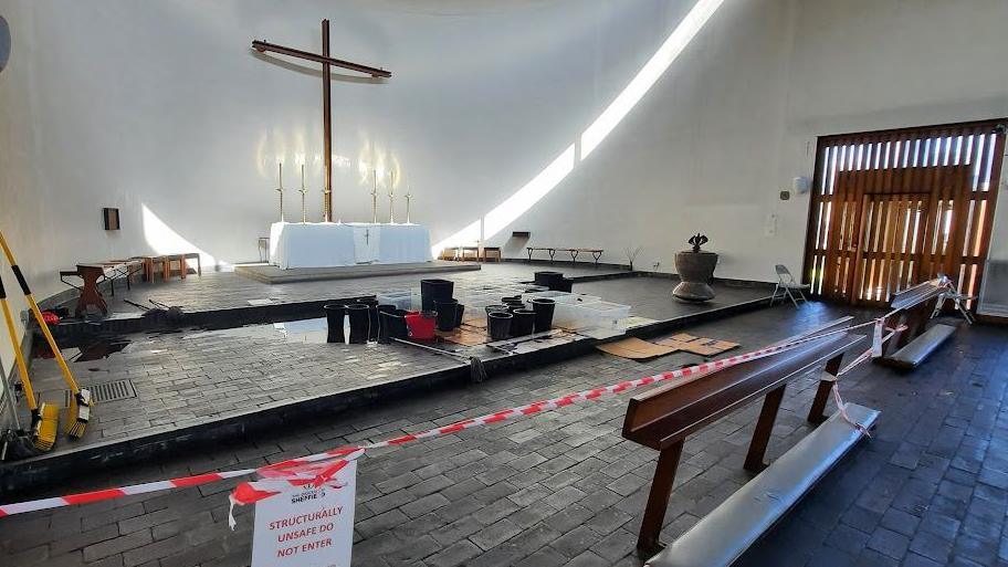 The front of a church. The floor is black and tiles. There are candles and a large cross against the front wall. There are brushes learning against walls and many buckets and containers sat on cardboard, presumably collecting water coming from above.