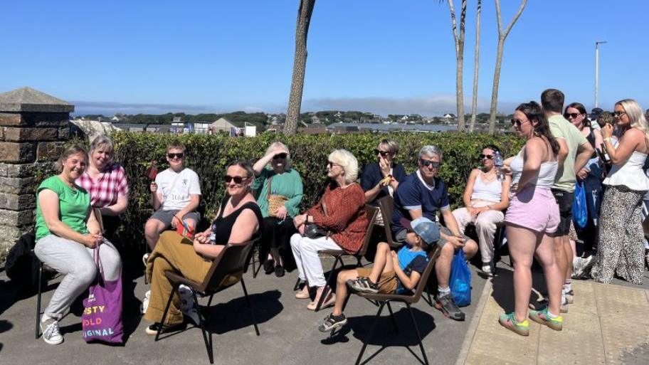 A queue of people sat outside a park with palm trees in the background.