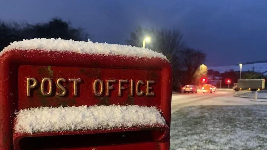 A red post box, with the words post office in gold writing, has snow sitting on top of it. It is next to a snow-covered road.