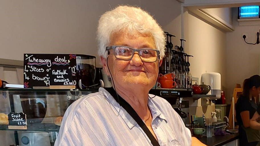 Margaret Peacock standing in a cafe smiling.  She has short white hair and grey framed glasses. She wearing a striped light-blue blouse.