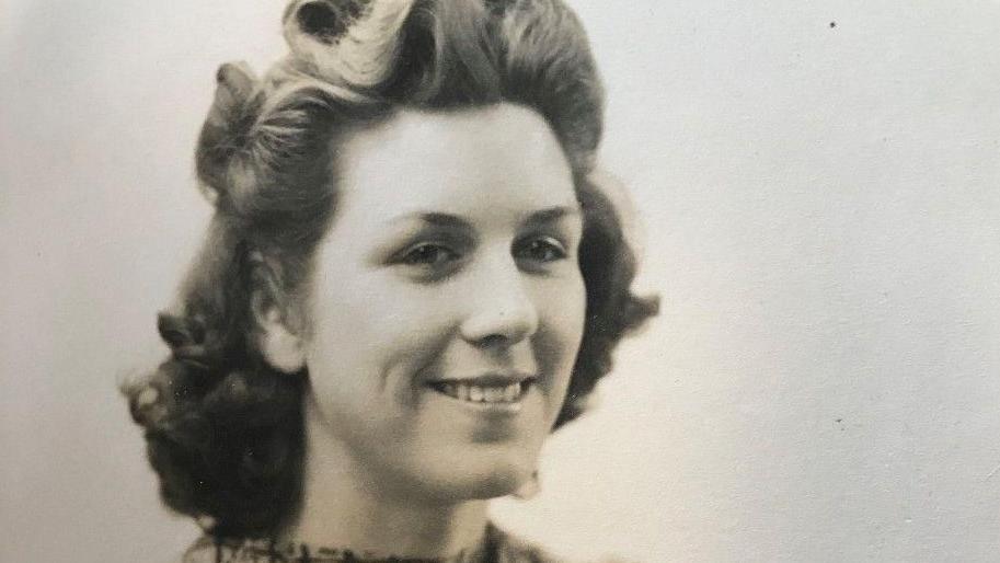 A black and white photograph of Mrs Bond when she was younger. She is wearing a high neck blouse with a square pattern print on it. She is smiling and glancing at the camera with her hair down but styled into position