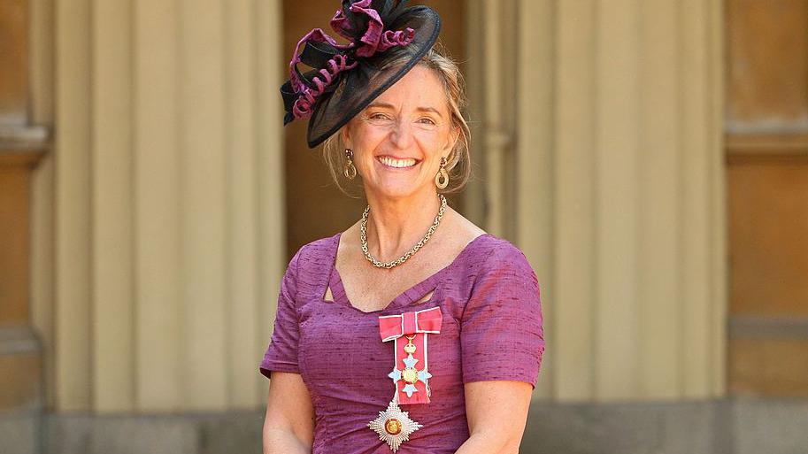 Dame Claire Bertschinger wearing a purple dress and a black hat. She has blonde hair and is wearing jewellery. She is smiling and looking at the camera.