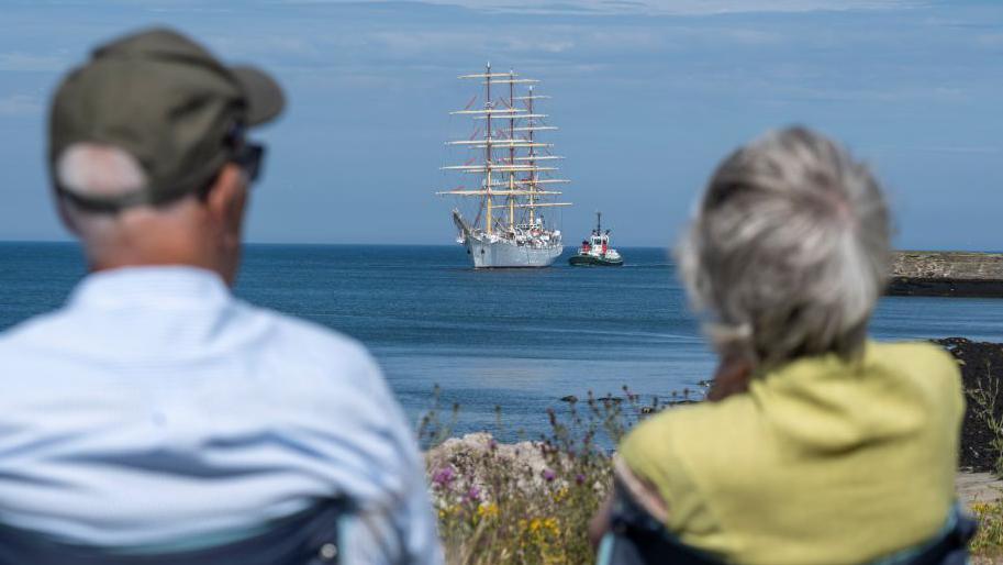 Tall ship in the distance, with the backs of a man and woman blurred in the foreground.