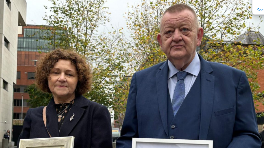 A woman and a man pictured outside a building. The woman is on the left, she is middle-aged with short brown curly hair and is wearing a blue jacket. The man on the right is older, with a blue suit, blue tie and blue shirt. 