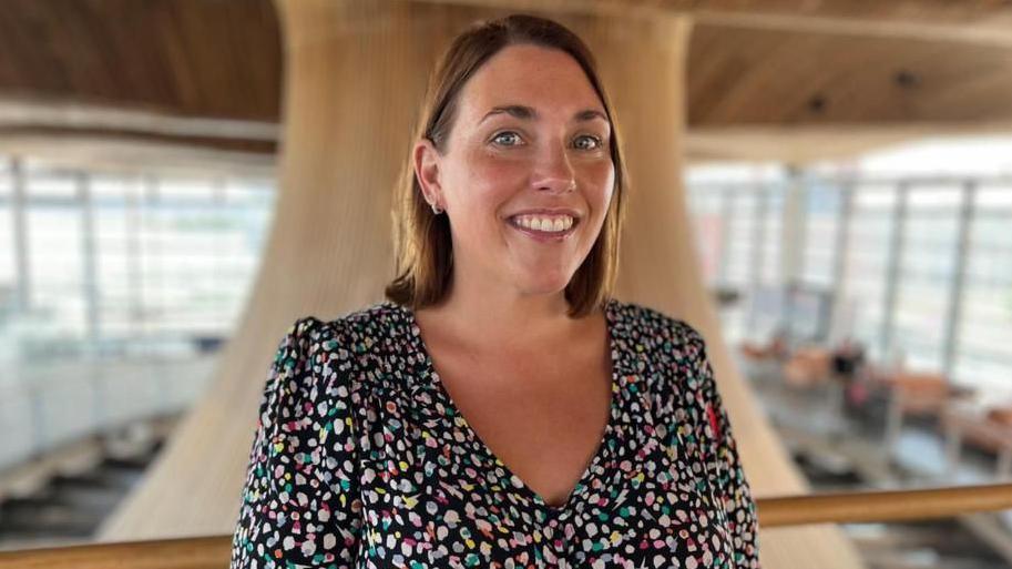 The image shows Heledd Fychan standing in the Senedd looking directly at the camera and smiling. She is wearing a colourful dress. The Senedd's funnel is behind her. It is a head and shoulders shot of her.