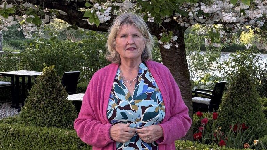 Carol Peet, founder of a buying agency, in a flowery dress, with a hedge and trees behind her