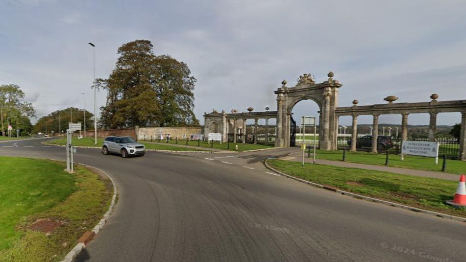 A roundabout on the A5 at Towcester Racecourse. A silver car is driving past a grand gate and a row of columns at the entrance of the course.