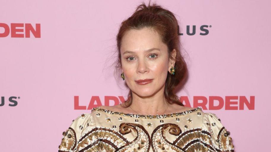 Anna Friel poses for a photo at an event. She has red hair, which is tied back in a high wavy ponytail. She has green gemstone earrings in and wears an off the shoulder embroidered and gemstone top, in beige, gold and brown colours. The background is a pale pink with the logo 'Lady Arden' in a deeper red colour.