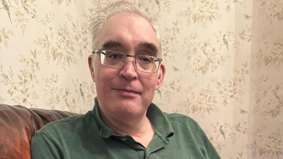 Andrew Lock is looking at the camera while sitting on a sofa in a living room, with light-coloured wallpaper with leaf and branch patterns behind him. He is wearing an olive green polo shirt and glasses. He has short grey hair.