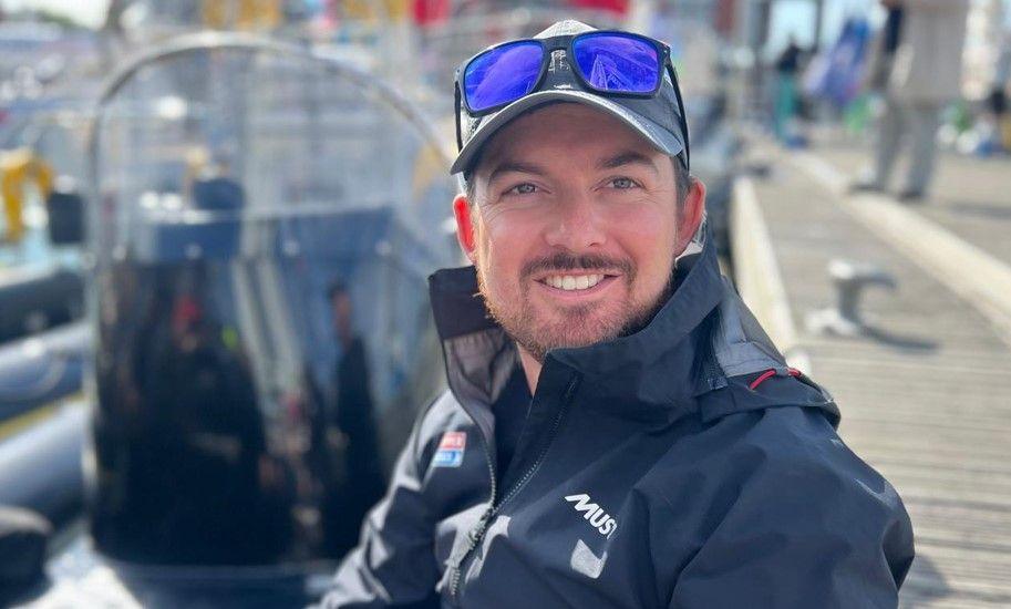 Max Rivers smiling for the camera. He's wearing a navy blue baseball cap with a pair of blue mirrored sunglasses resting on the peak. He has a full dark brow beard and is wearing a navy blue waterproof jacket.