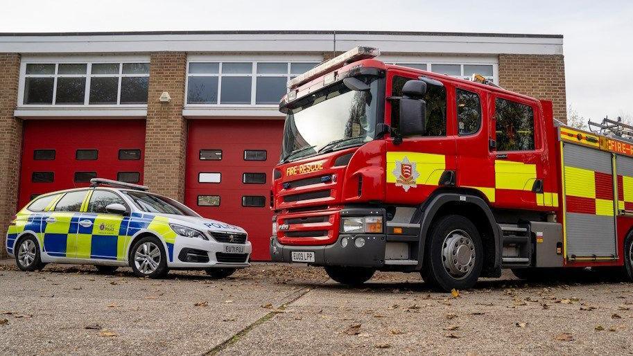 A red and yellow fire engine parked next to a blue, yellow and white police car. They are both in front of a fire station, which has large red doors.