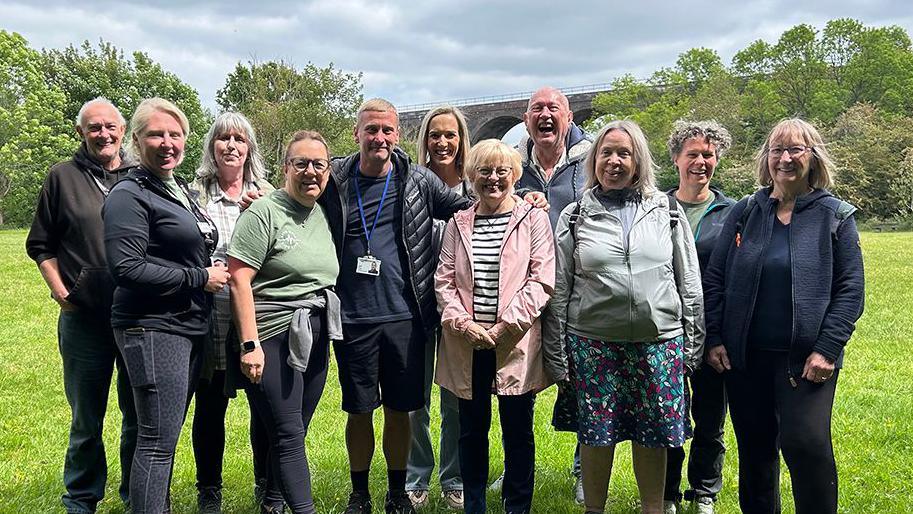 A group of walkers are pictured in a field in Reddish Valley Park, Stockport. They are grouped closely together and are all smiling on a sunny day.