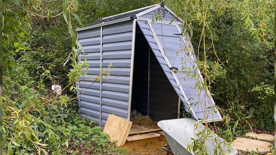 The front door of a small storage shed is partially ripped off.