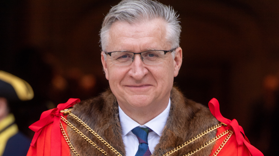 Alistair King wearing a red cloak lined with brown fur with gold chains over the top. He has grey hair and is wearing glasses