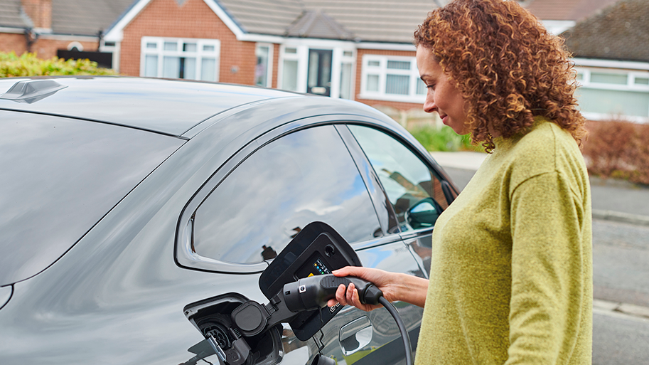 A woman in a yellow/green jumper inserting an electric vehicle charging cable into a dark-coloured coupe-style car. There are some bungalows in the background