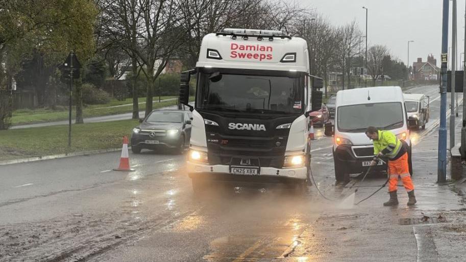 A lorry in a flooded street with a car passing by on the other side of the road, past an orange traffic cone, with a man with a hose attempting to clean the roadside