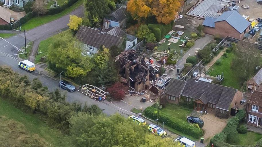 An aerial view of a house which has been destroyed by a fire. There are emergency vehicles on the road alongside the house.