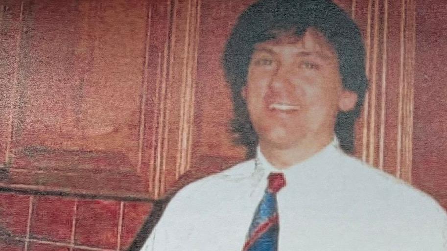 Archive photograph of Ricky Haywood smiling and wearing a white shirt and blue and red tie. He is standing in front of wood cupboards