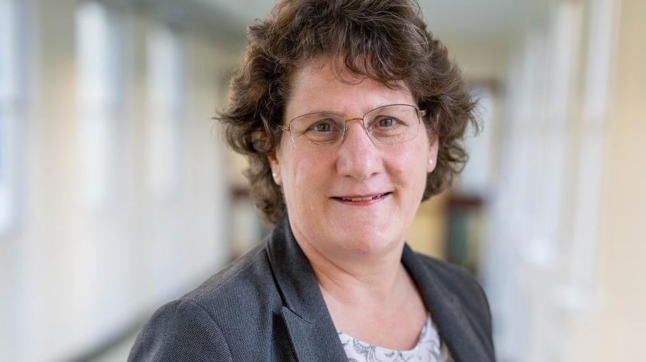 A woman with short curling dark hair wearing wire framed glasses and a dark grey jacket, looking towards the camera and slightly smiling