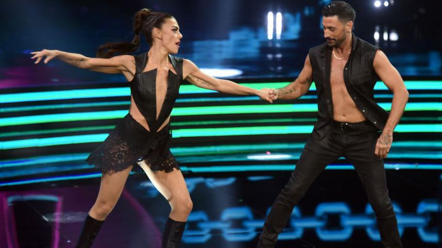 Bianca Guaccero and Giovanni Pernice dressed in black outfits hold hands mid-dance routine with green and blue neon lights in the background.