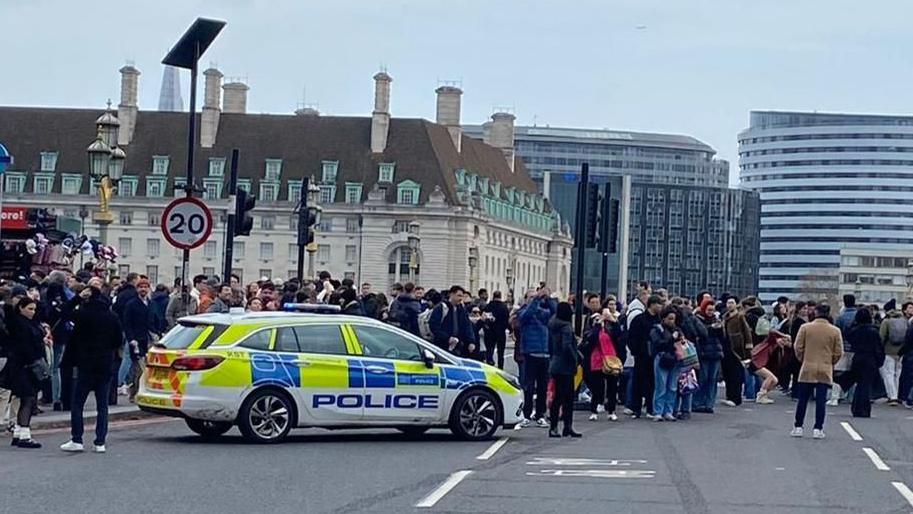 A police car has stopped on Westminster Bridge and people are stood all around it in the background