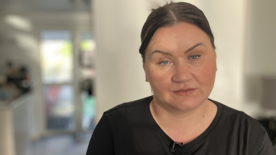 Michelle looks at the camera with a serious expression. She has dark hair tied back in a centre-part, and wears a black t-shirt.