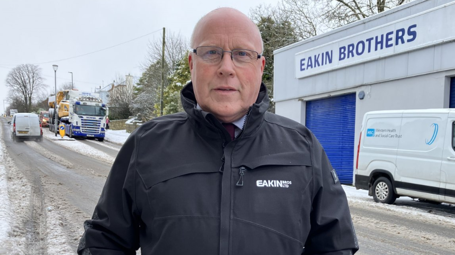 Shows Peter Eakin wearing a company coat, standing on the roadside with traffic in the background