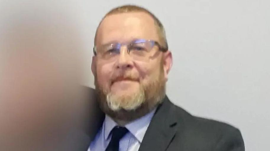 A man with very short hair and light facial hair. He is wearing glasses and a suit, and is looking into the camera. He is stood in front of a grey background