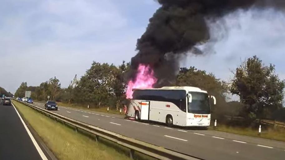 Dashcam footage taken from the other side of the dual carriageway. The white coach has pulled over in the layby and red flames have engulfed the rear section of the vehicle. Large plumes of thick black smoke are billowing in the area.