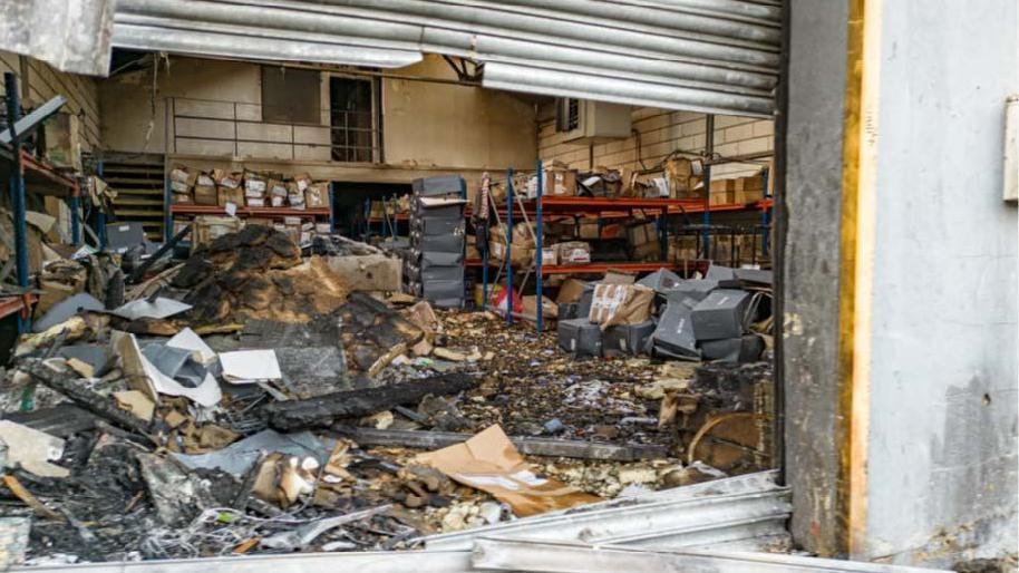An image underneath a metal shutter shows a burnt out warehouse