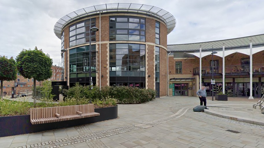 A modern building in a circular shape in brick and glass with trees and green spaces outside and benches and paved walkways