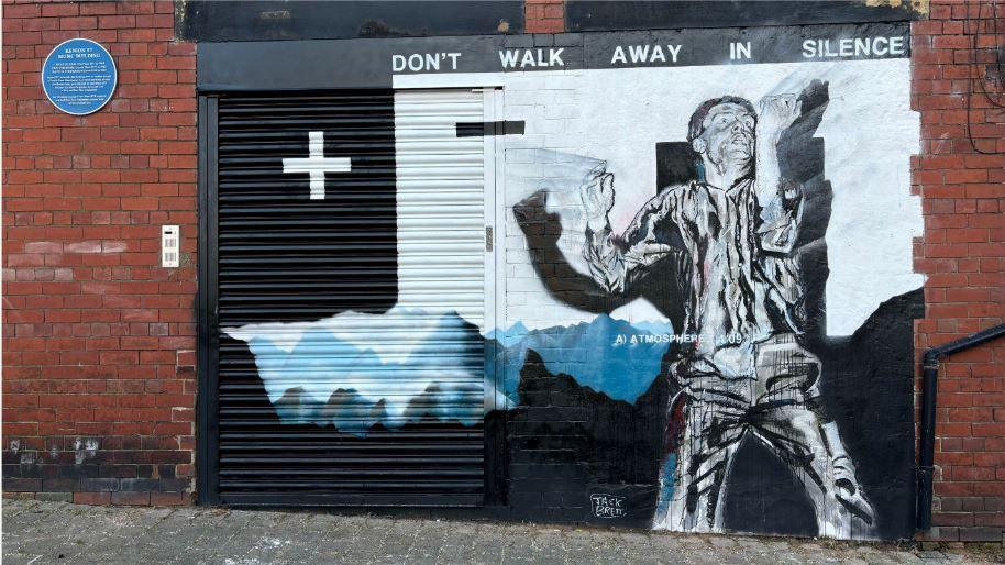 A black and white mural of Ian Curtis, stood on stage holding up a microphone, painted on to a redbrick building, over a doorway, with the lyrics 'don't walk away in silence' painted above.