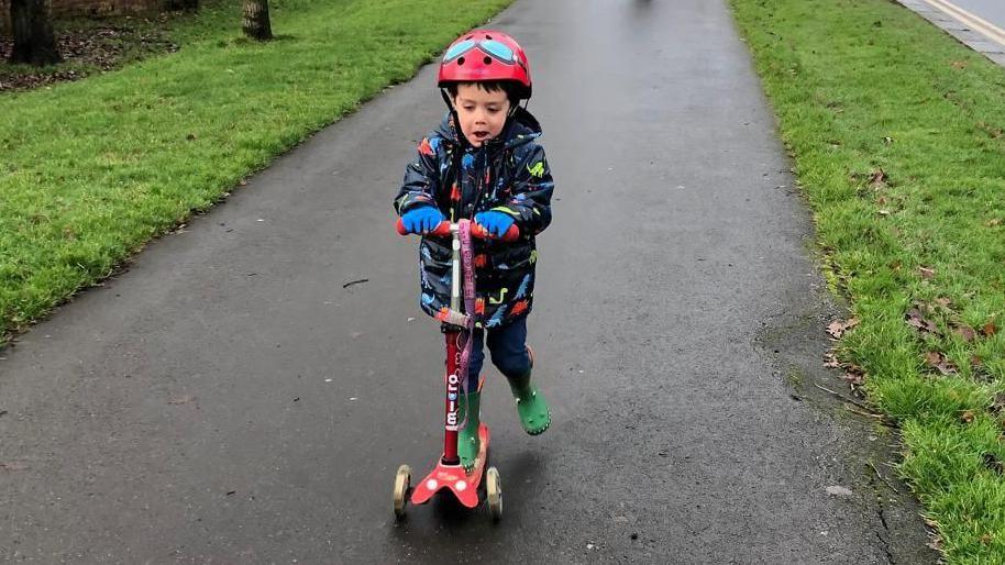 Sam Liew riding a red scooter, wearing a red helmet, a coat with dinosaurs on, blue gloves and a red helmet