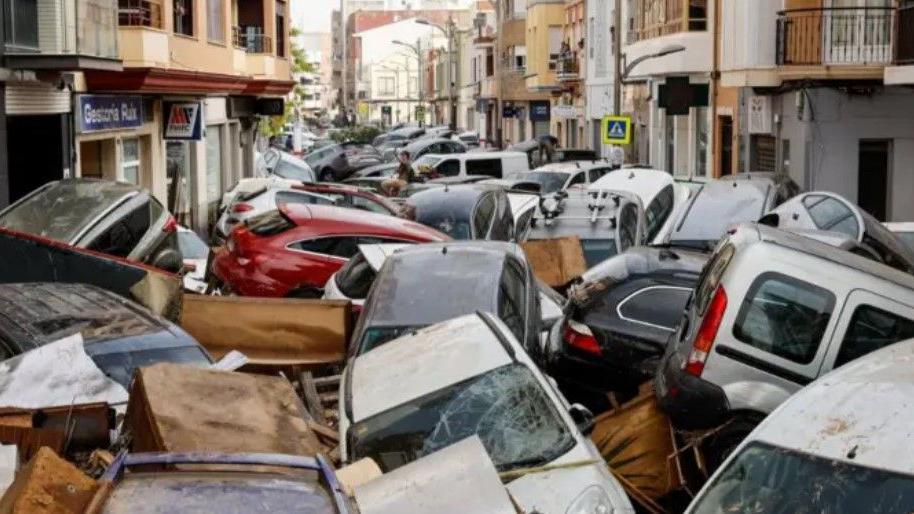 Cars piling on top of each other in Spain. Some have smashed windscreen and they are dotted as far as the eye can see on a street with houses on each side.