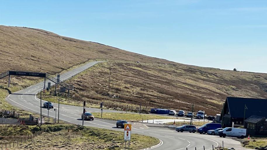 The Mountain Road stretching from the Bungalow through to Brandywell. It is on a sloping mountainside covered in heathland. There is a building at the Bungalow on the right with cars parked in front of it and there are three cars travelling on the road. There is a metal footbridge over the road on the left.