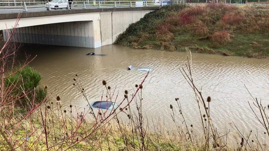 A road under a bridge is completely flooded, with the roofs of a grey and black car just about visible over the water's surface