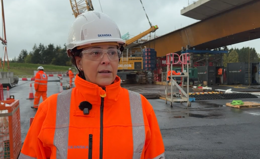 A screenshot of a video interview with Rosario Barcena at the site where the bridge is being built. She is wearing a white hard hat, safety goggles and orange high-viz jacket. She is looking in the distance while being interviewed by someone out of shot. She has a microphone pinned to the side of her jacket. Behind her are heavy plant machinery and workmen in high-viz clothing working around the steel structure of the new bridge.