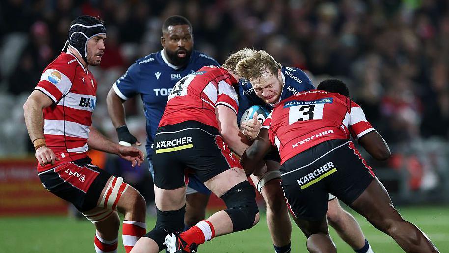 Two Gloucester players tackle Ben Bamber of Sale as he bends forward with the ball in his hands and his eyes closed. A Gloucester and Sale player stand behind and to the side