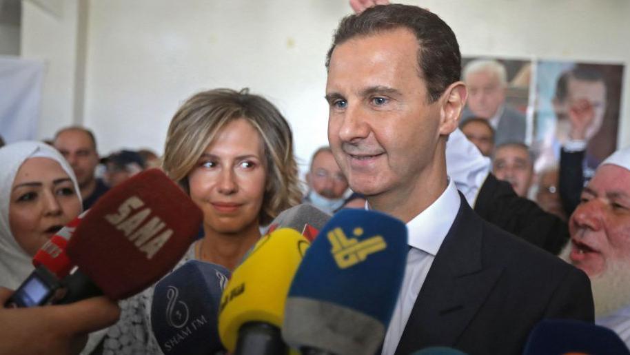 Microphones partially block a view of Asma al-Assad, smiling at her husband, who stands in front of her addressing the microphones. 