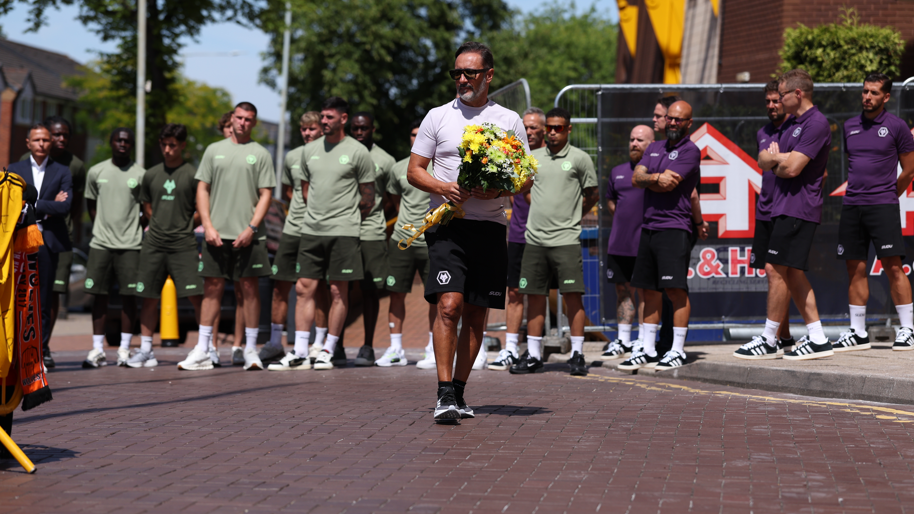 A man in a white t-shirt and black shorts holds a large bunch of flowers as he walks along a pavement. There is a row of men in football training tops behind him.
