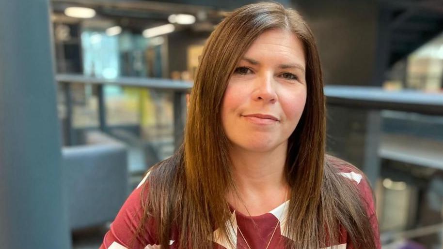 A head and shoulders photograph of Laura Doel at BBC Wales' Cardiff headquarters. She has long brown hair, is wearing a red and white top and looking at the camera. There is the back of a large blue chair and steps leading up to the floor she is on in the blurred background behind her.