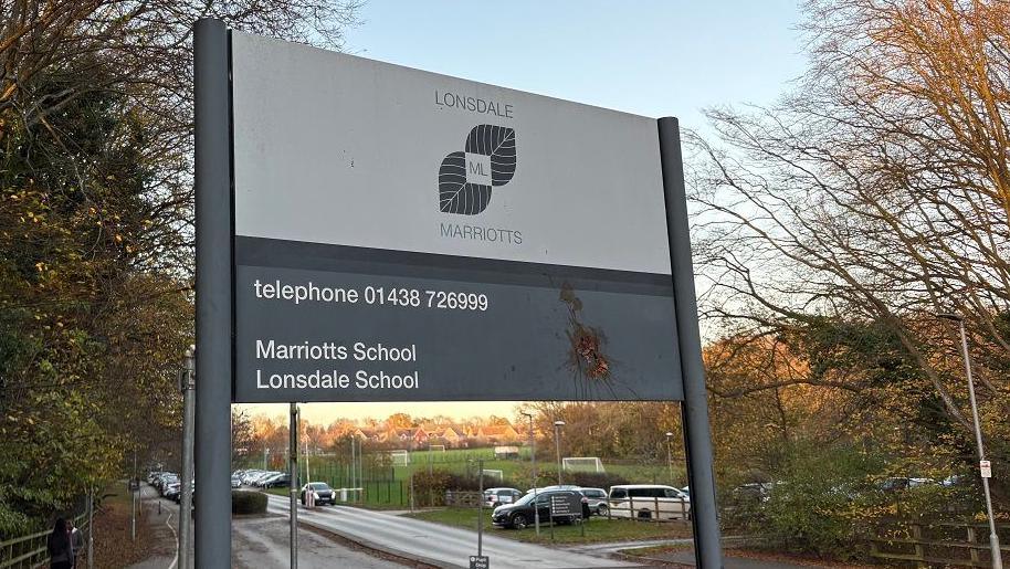 A sign at the entrance to school with "Lonsdale School" in grey lettering with a grey leaf logo. There is also a telephone number in white lettering on a grey background. There are trees on both sides of the sign and a car park beyond it.