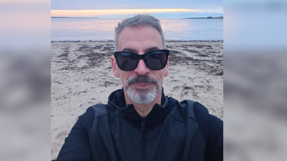 A man wearing black sunglasses, sporting grey hair and a grey french beard looks into the camera while standing on a beach with the sea behind him.