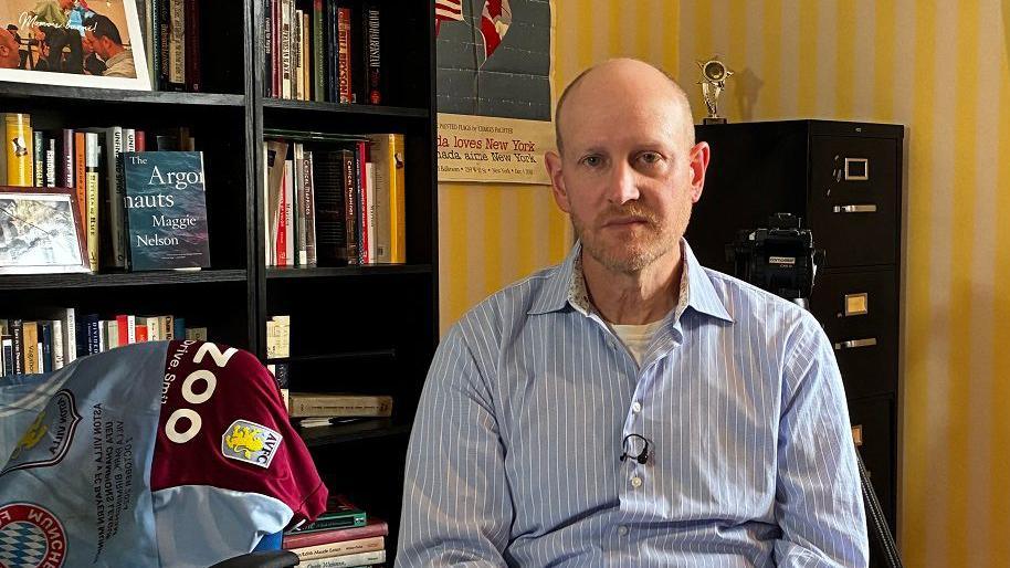 Elliot Ludvig sits at home with a bookcase and pictures behind him. He is wearing a light blue shirt and his Villa fan shirt is next to him on a chair. He is looking at the camera with a concerned expression.