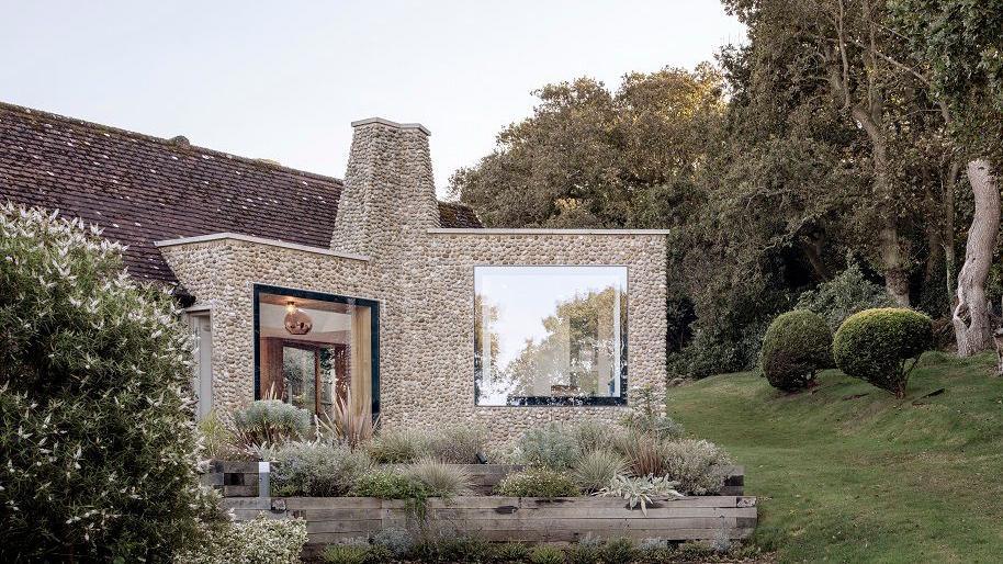 Single-storey building made from small grey stones. It has two square sections with a pointed chimney between. There are picture windows on each section. There are shrubs in front of the building and grass to the right with circular bushes.  There are further bushes to the right and left.