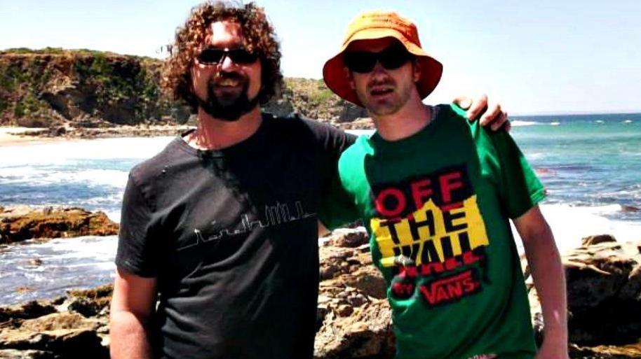 Two men standing next to each other on a beach which has rocks, the sea and the base of a cliff showing. The man on the left has curly hair and a beard and is wearing a black t-shirt with a curvy line design on it. The man on the right has a green t-shirt which says '"Off the Wall" in black letters against a yellow rectangle.