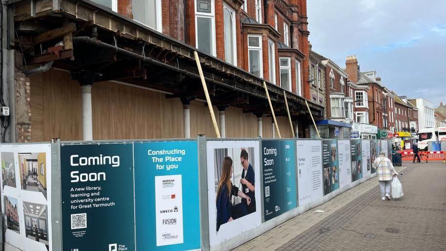 Hoardings surround the facade of the old Palmers Department Store with wooden boards behind the iron pillars, and canopy above, and brickwork with bay windows above that. A man carrying a bag is walking down the pedestrianised King Street.